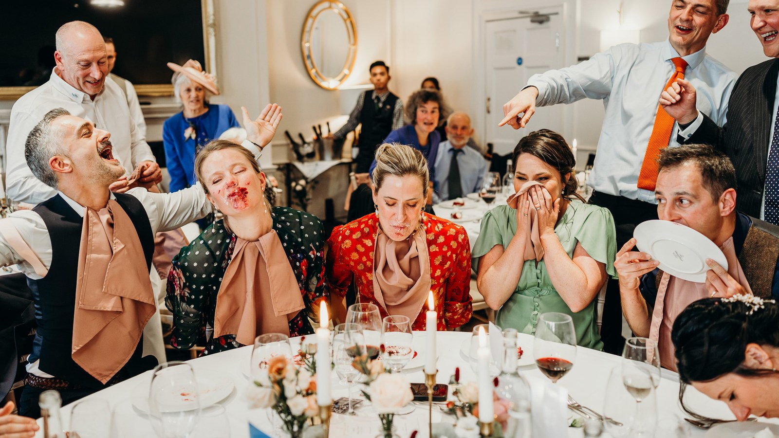 Guests at a wedding reception table are mid-action during a game, with several covered in cake or