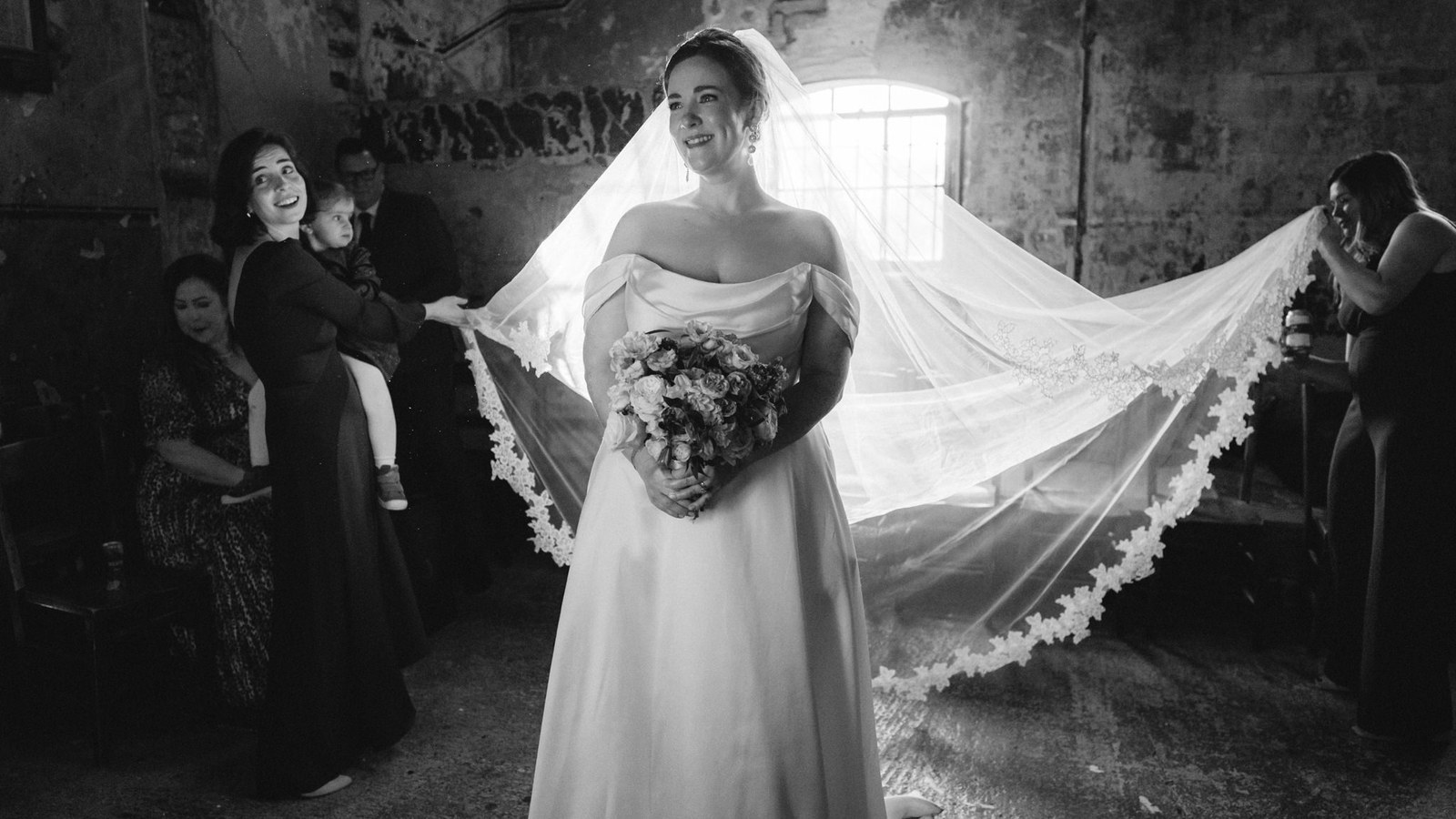 A bride stands smiling, holding a bouquet, with her long veil fanned out behind her, held by