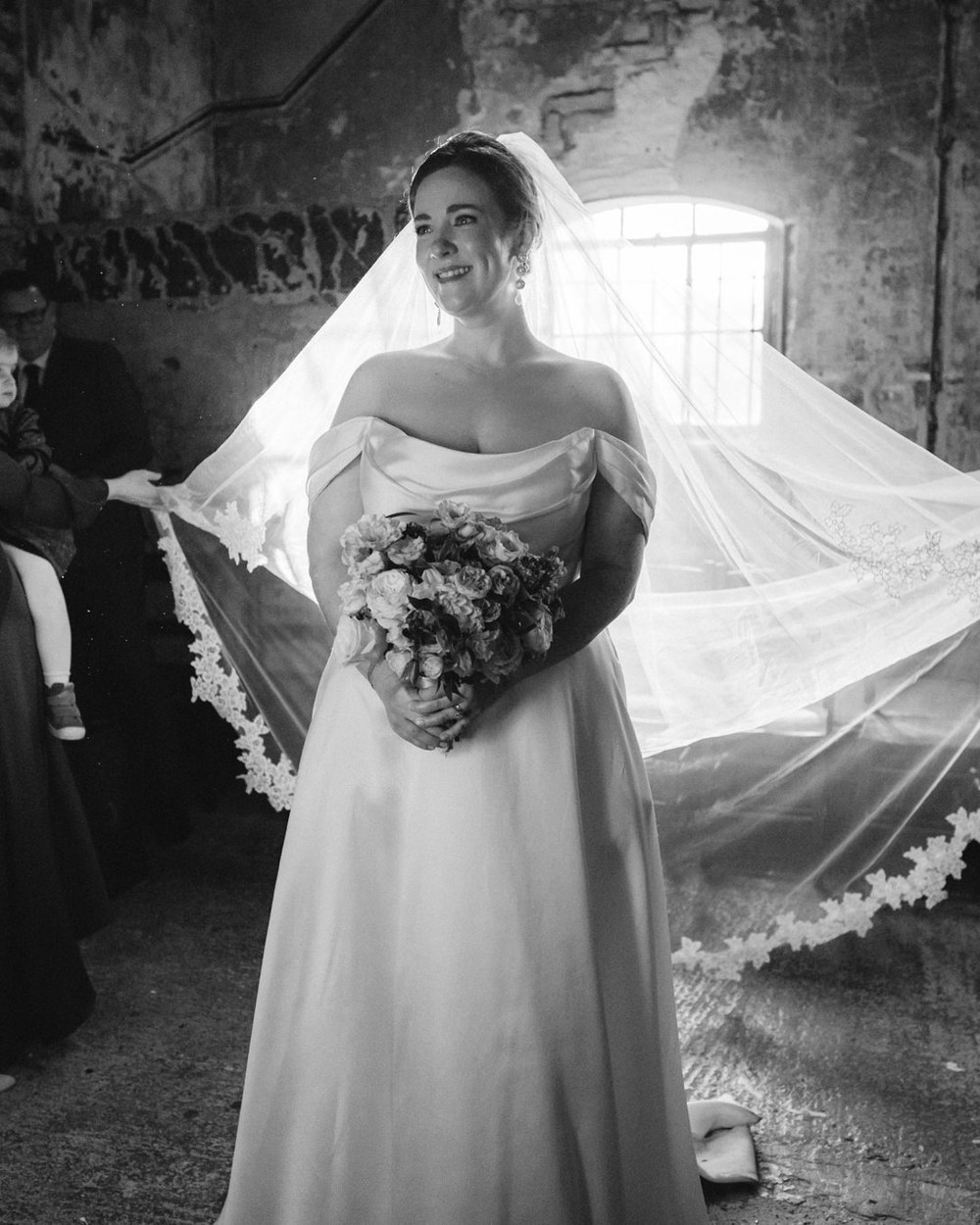 A bride stands smiling, holding a bouquet, with her long veil fanned out behind her, held by