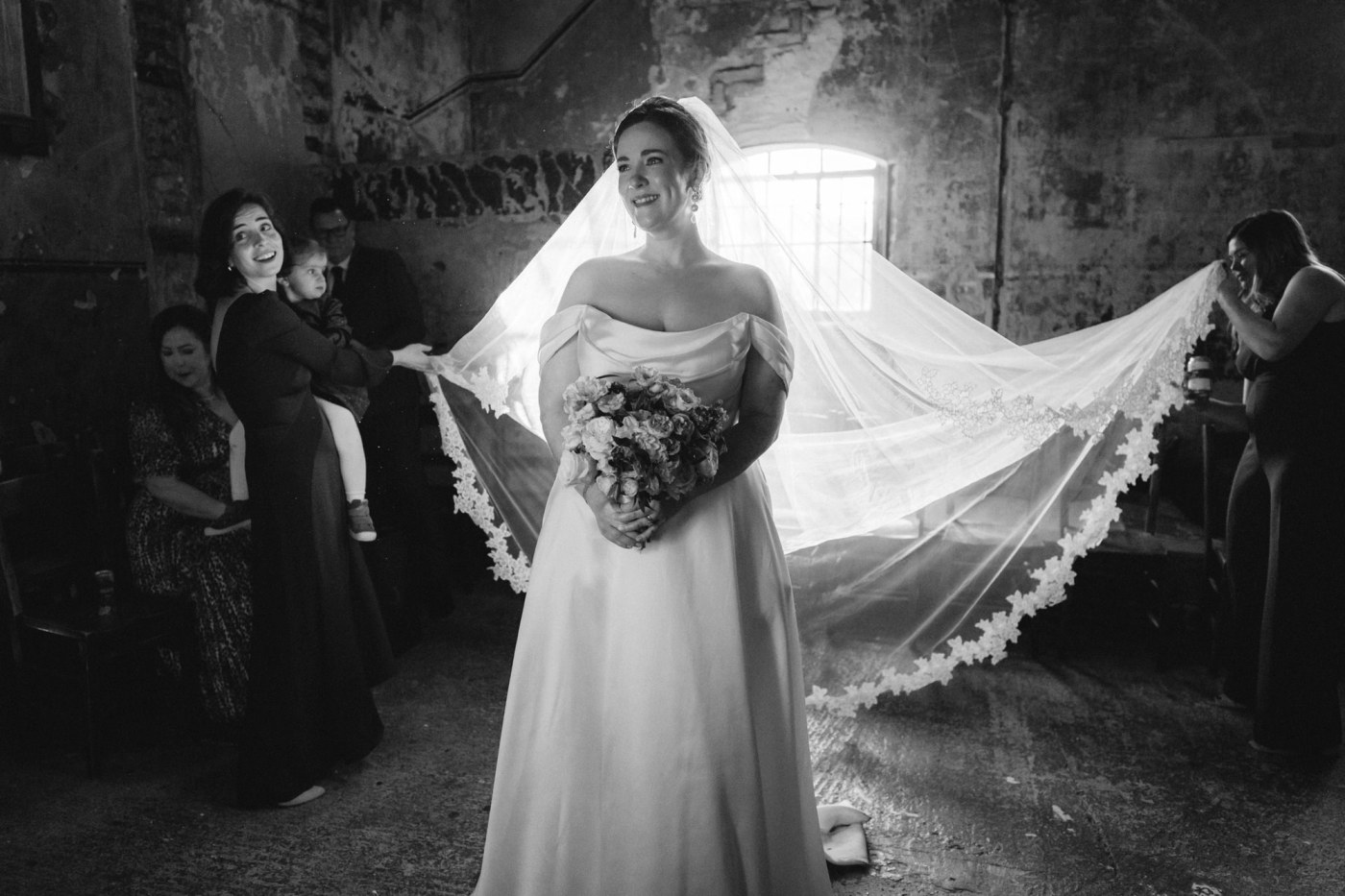 A bride stands smiling, holding a bouquet, with her long veil fanned out behind her, held by