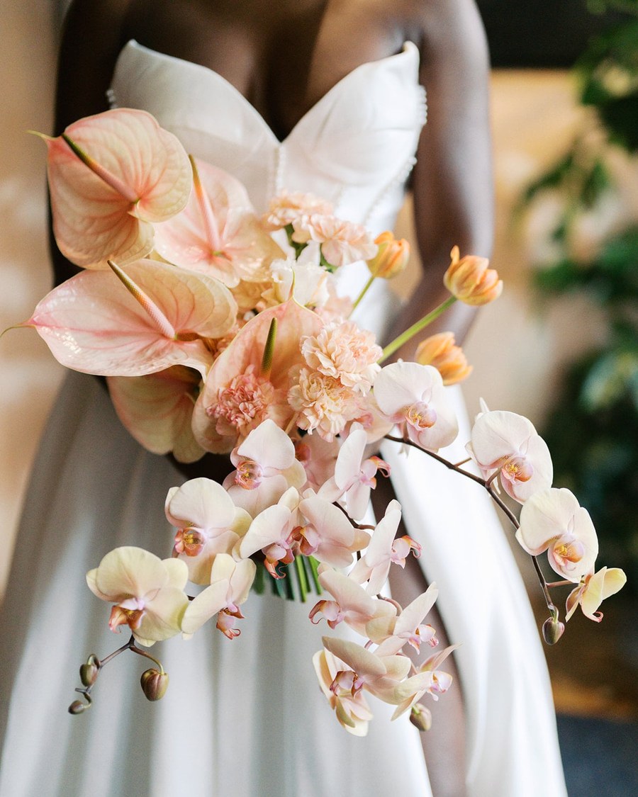 A bride holds an elongated, cascading bouquet featuring pale peach anthuriums, peachy-pink