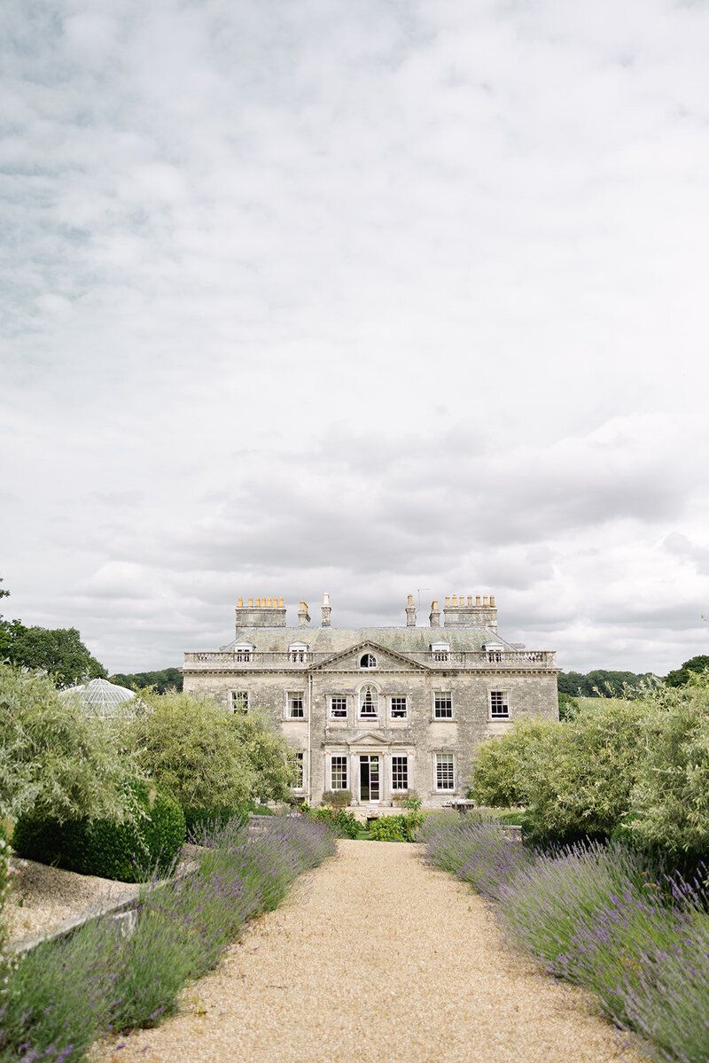 A grand, grey stone Georgian mansion with multiple windows and prominent chimneys stands beneath an
