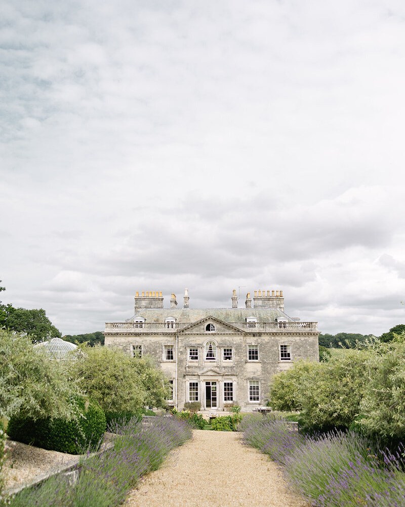 A grand, grey stone Georgian mansion with multiple windows and prominent chimneys stands beneath an