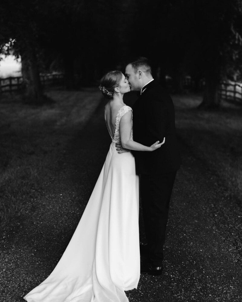 A bride in a white gown with an illusion lace back and a groom in a black suit share a tender kiss