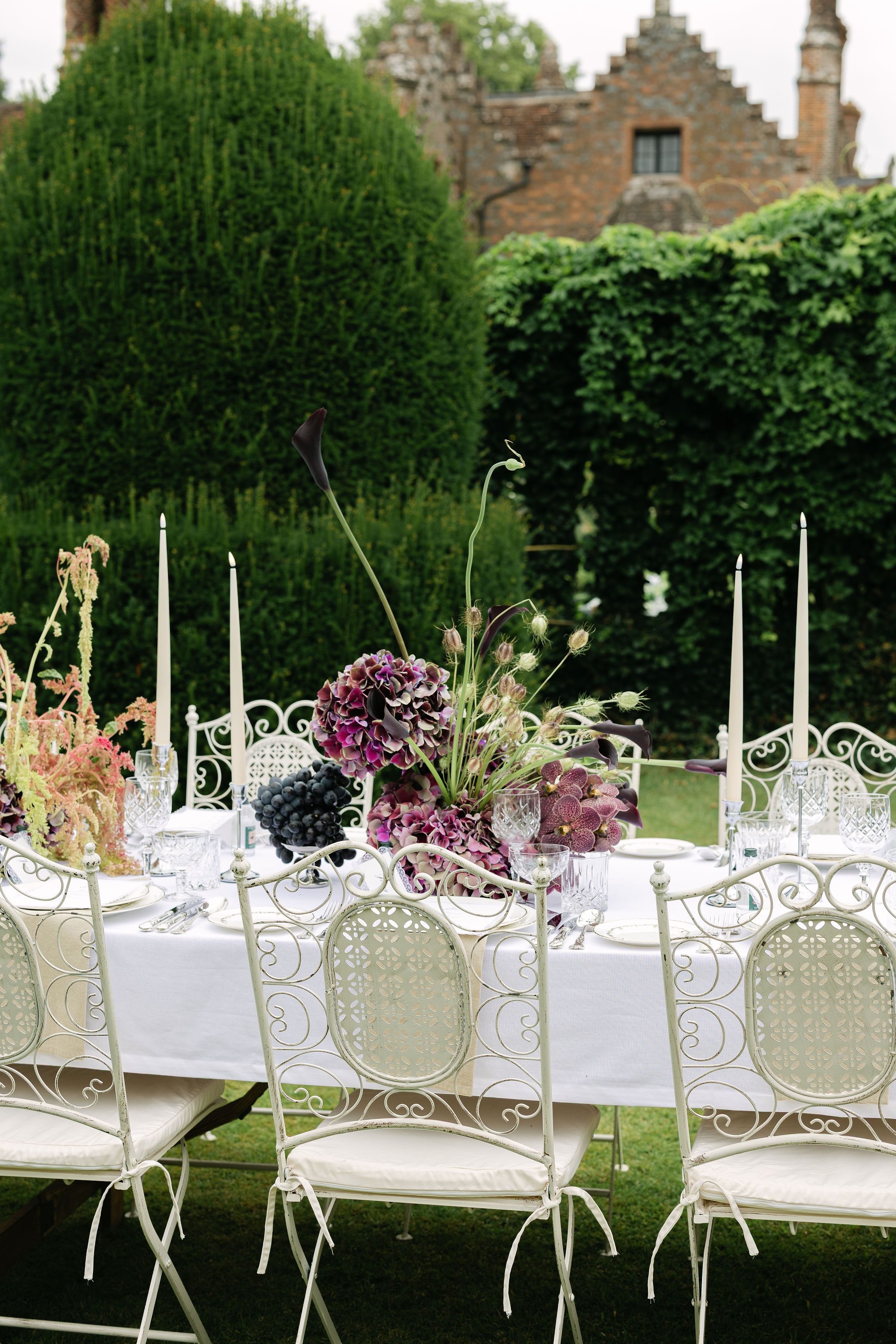 An outdoor wedding reception table is set with a floral centrepiece featuring dark purple