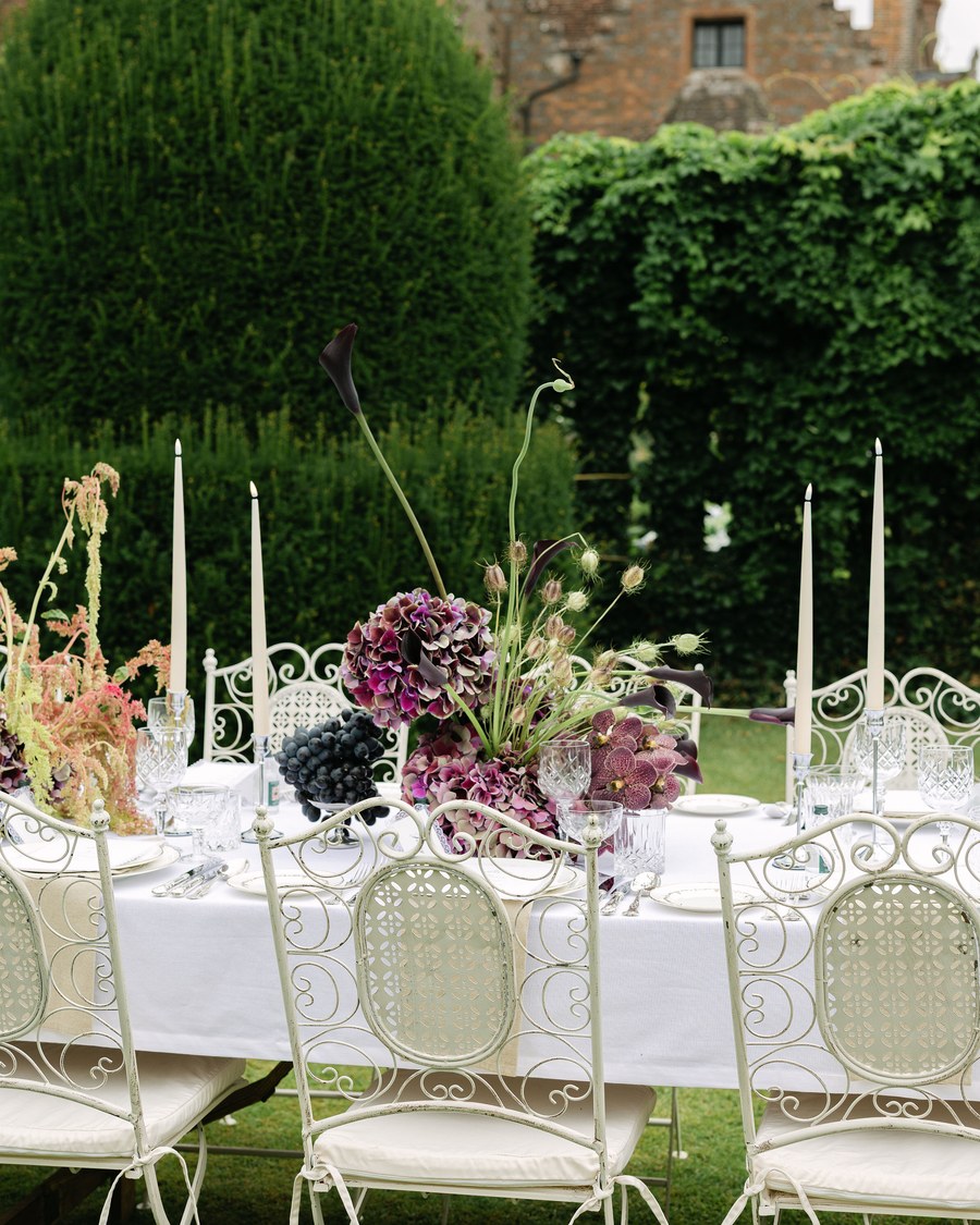 An outdoor wedding reception table is set with a floral centrepiece featuring dark purple