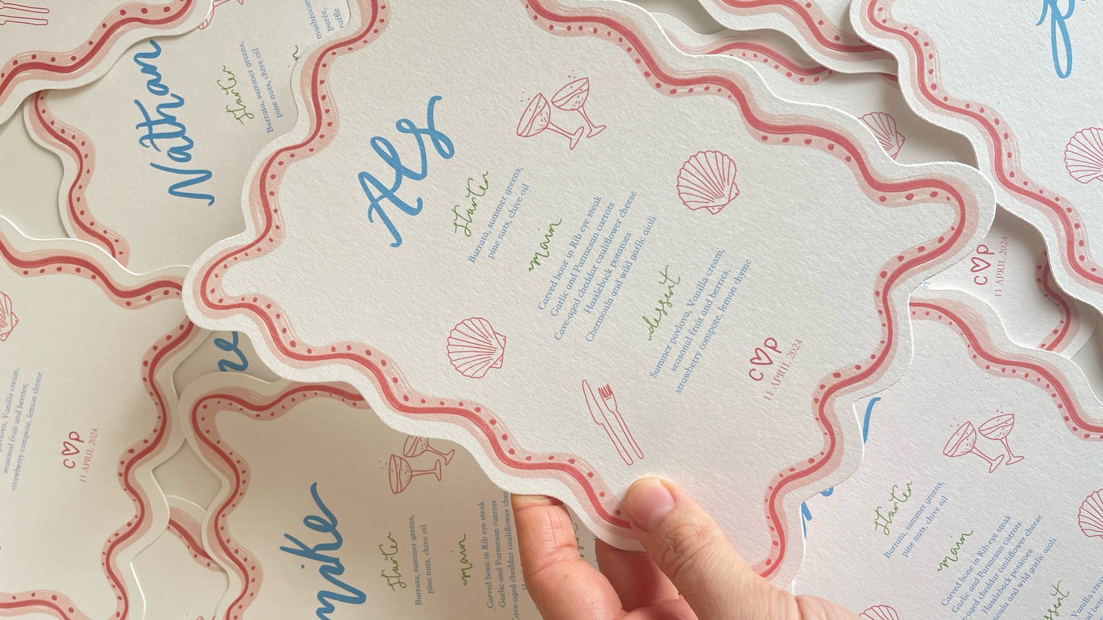 A hand lifting one wavy-edged wedding menu card from a stack, each printed with a guest's name in blue script above a three-course menu, edged in pink with small shell and cocktail-glass motifs.