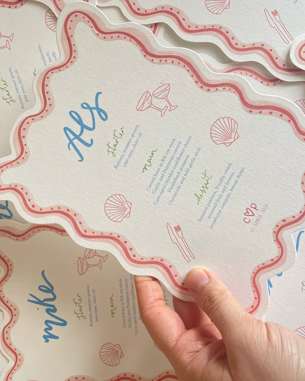 A hand lifting one wavy-edged wedding menu card from a stack, each printed with a guest's name in blue script above a three-course menu, edged in pink with small shell and cocktail-glass motifs.