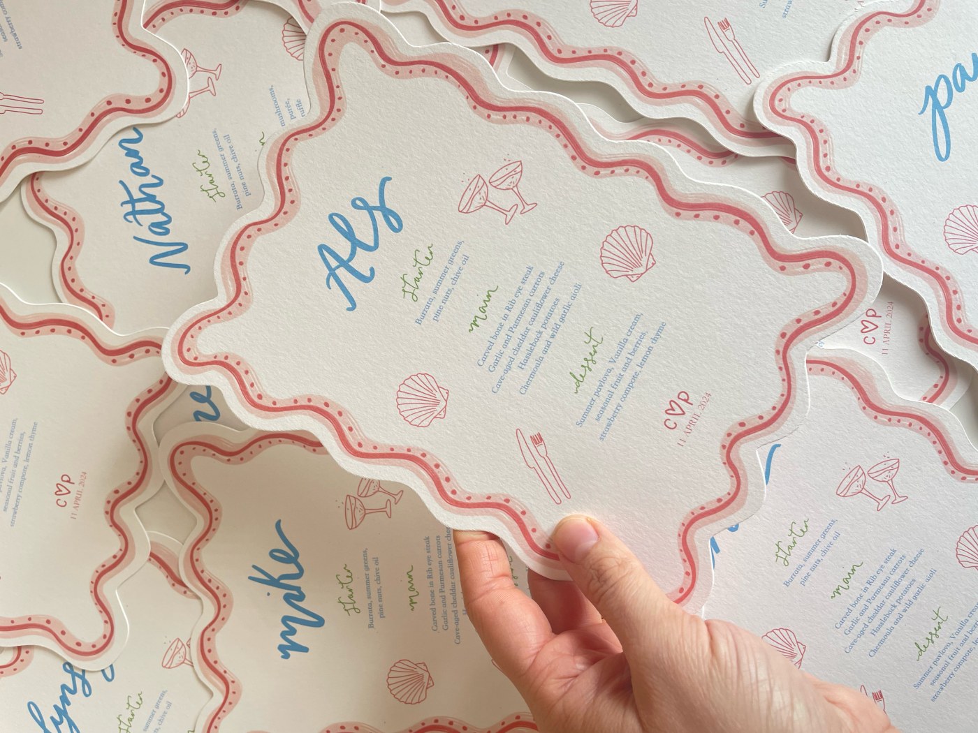 A hand lifting one wavy-edged wedding menu card from a stack, each printed with a guest's name in blue script above a three-course menu, edged in pink with small shell and cocktail-glass motifs.