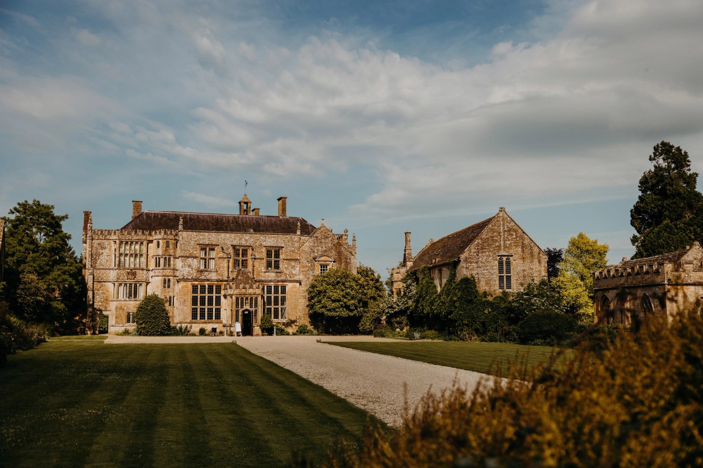 A wide exterior shot of a large, historic manor house made of light brown stone, featuring