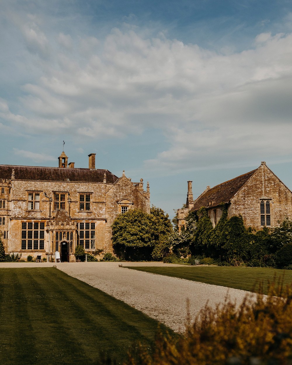 A wide exterior shot of a large, historic manor house made of light brown stone, featuring