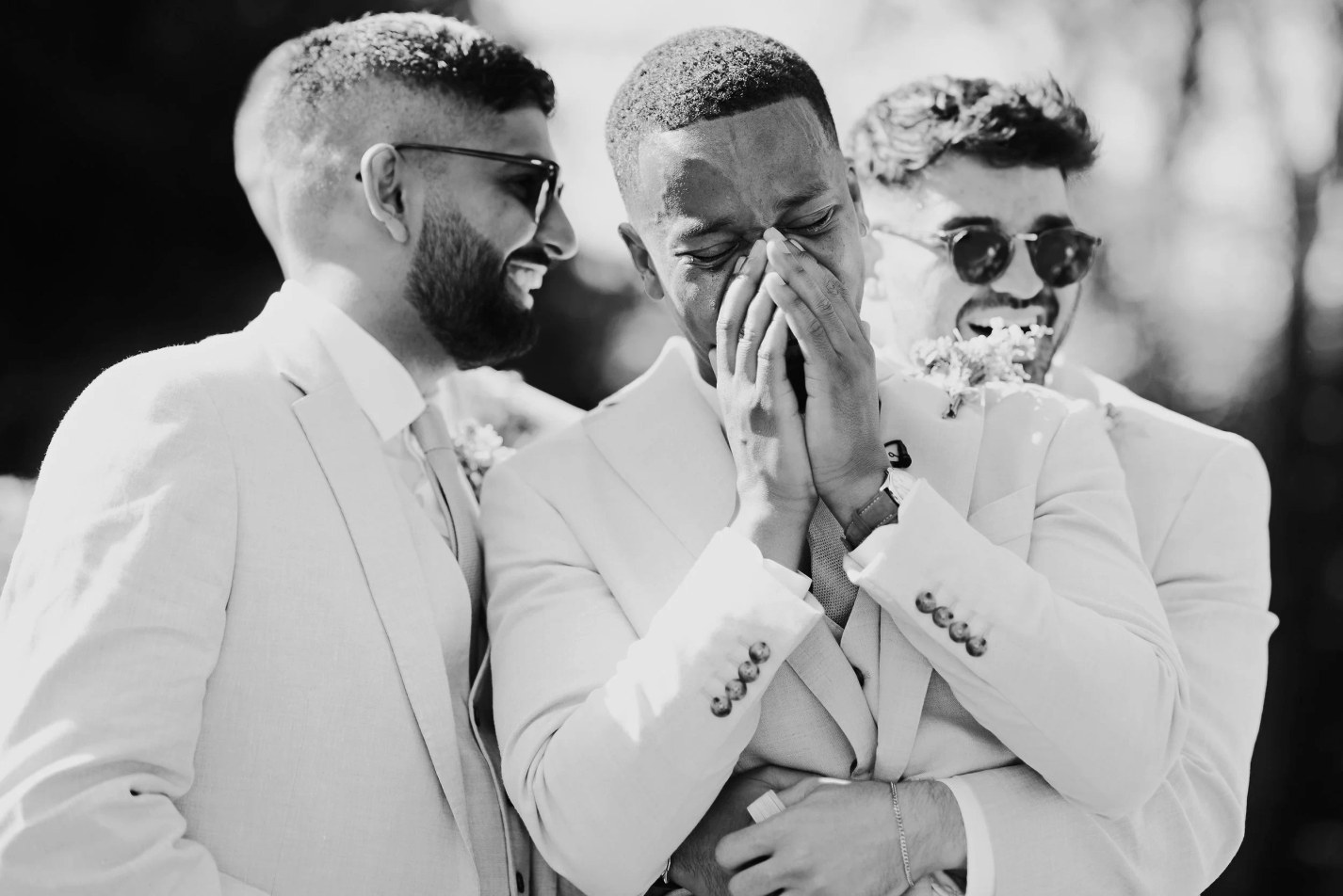 Three men, including a groom or groomsman, are captured in a candid black and white outdoor moment.