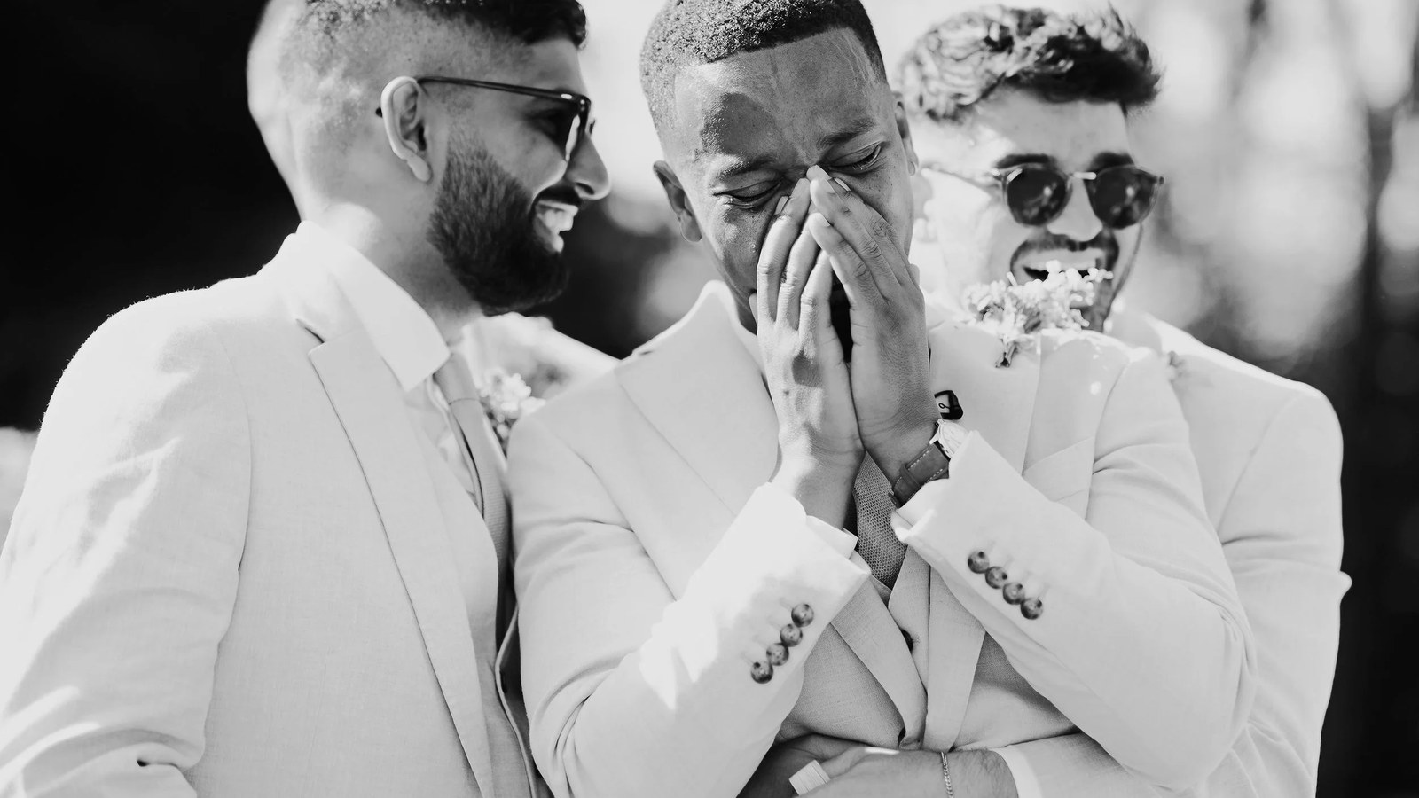 Three men, including a groom or groomsman, are captured in a candid black and white outdoor moment.