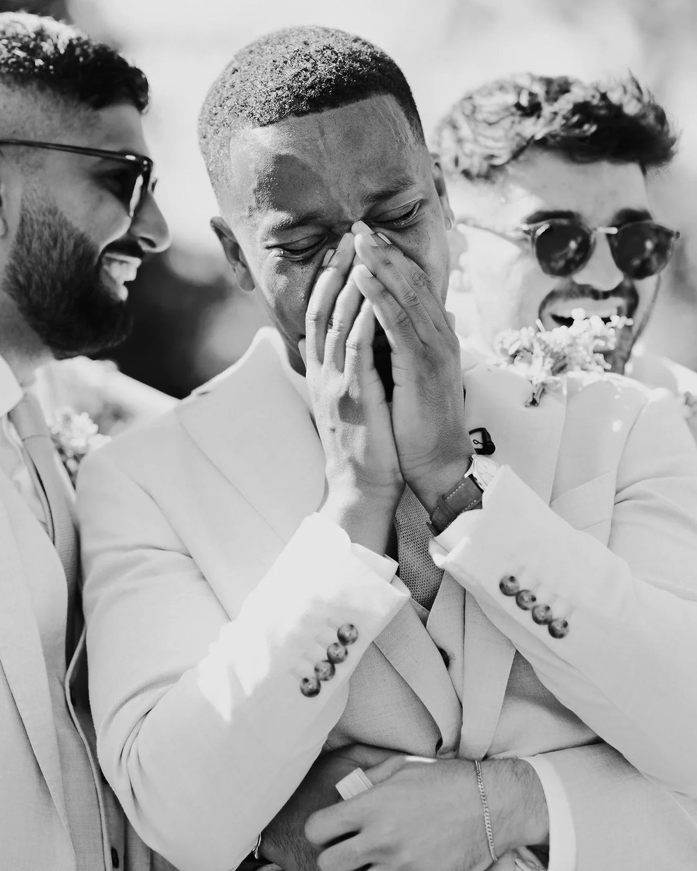 Three men, including a groom or groomsman, are captured in a candid black and white outdoor moment.