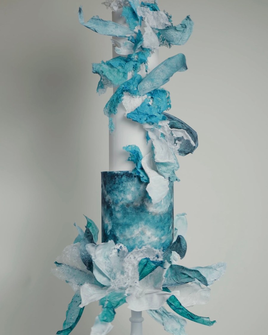 A three-tier round cake featuring a lower tier with a hand-painted blue and turquoise watercolour
