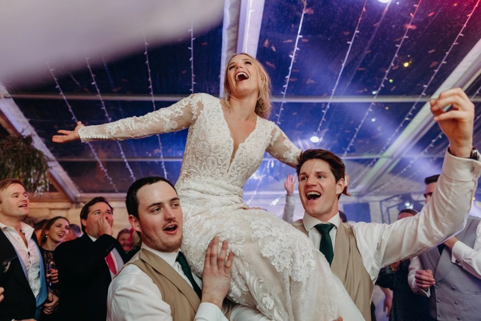 A bride in a lace wedding dress is held aloft on the shoulders of two men, laughing with her arms
