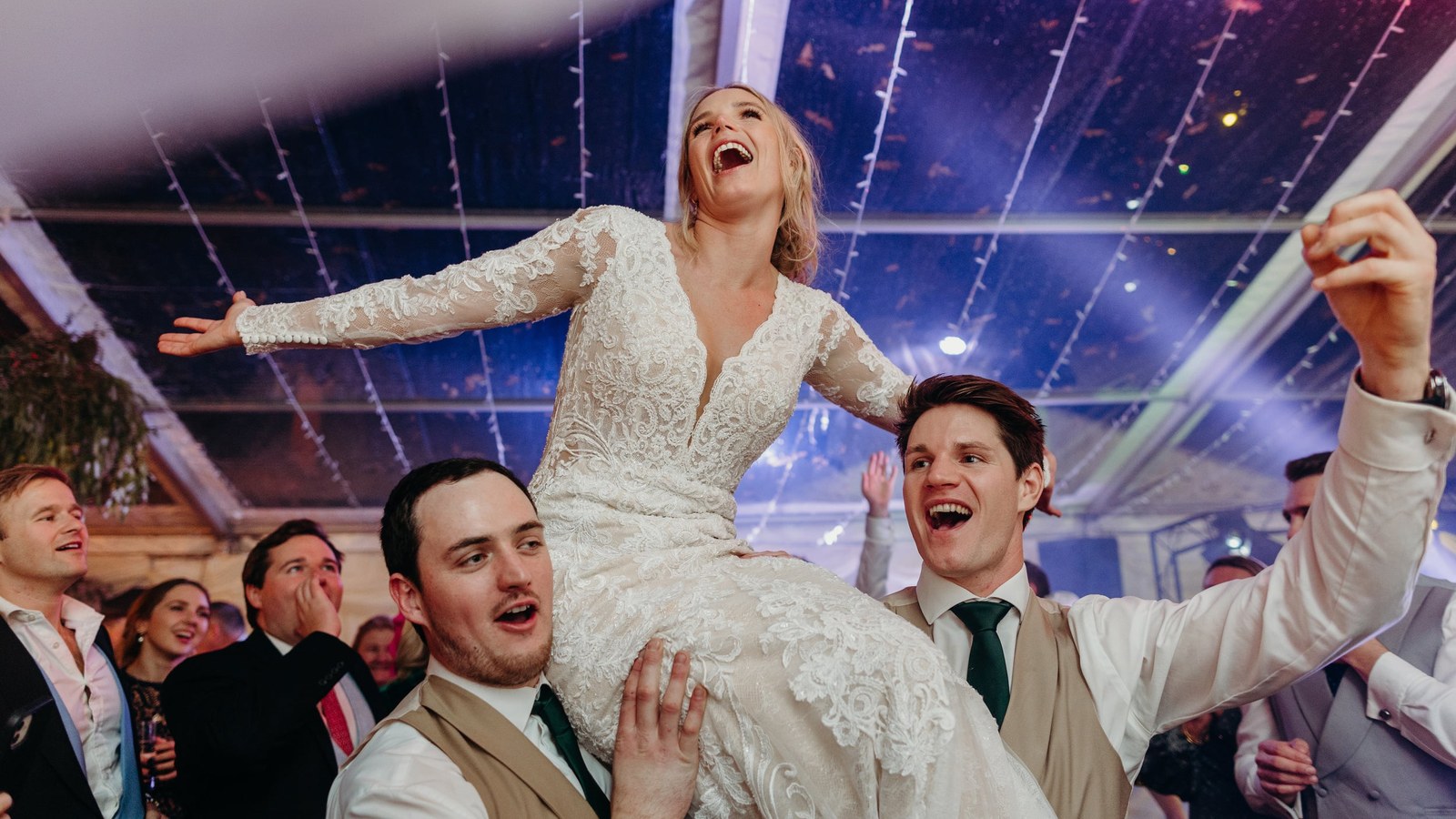 A bride in a lace wedding dress is held aloft on the shoulders of two men, laughing with her arms