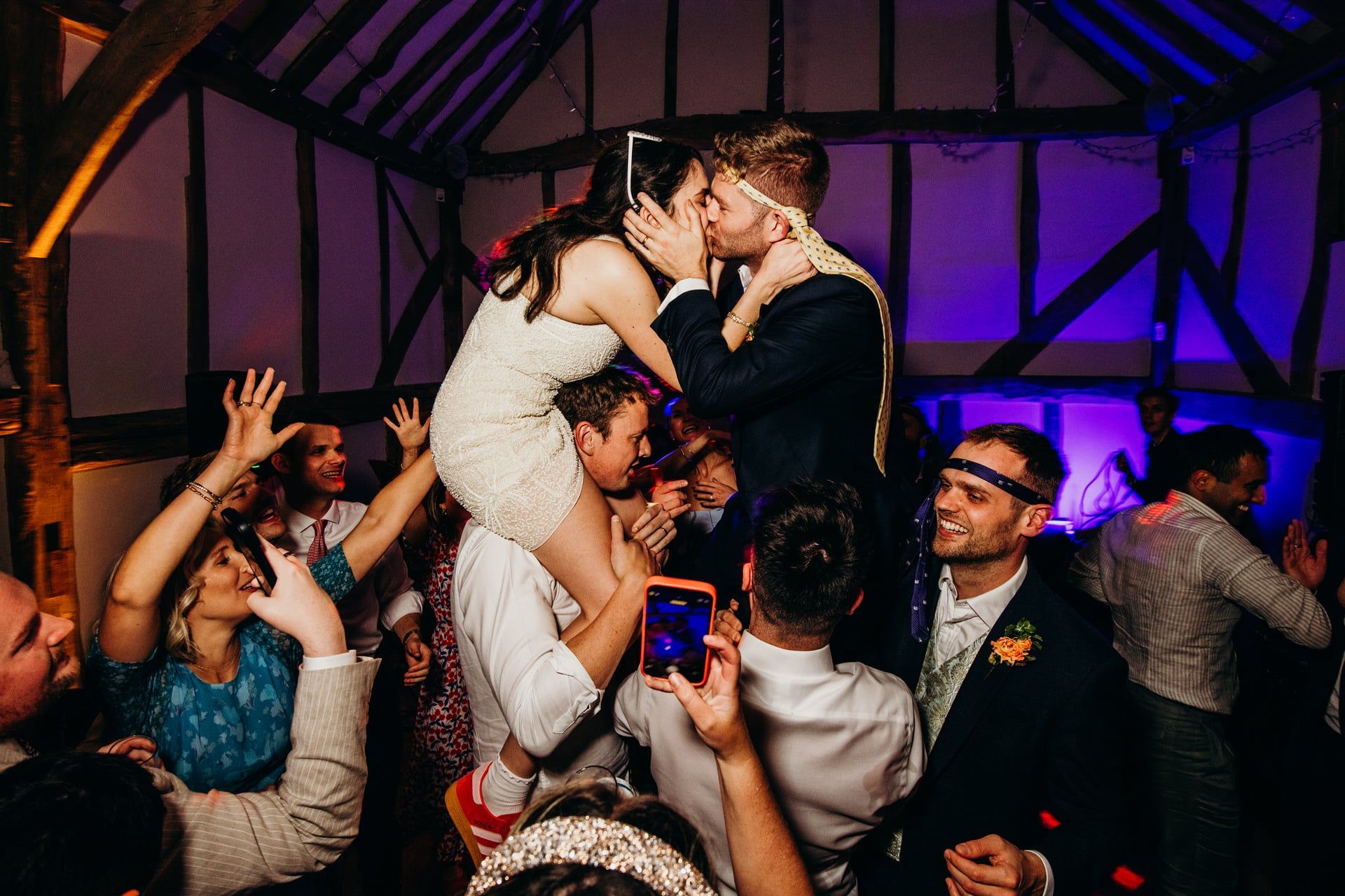 A bride in a short dress sits on a guest's shoulders, kissing the groom who is also on another