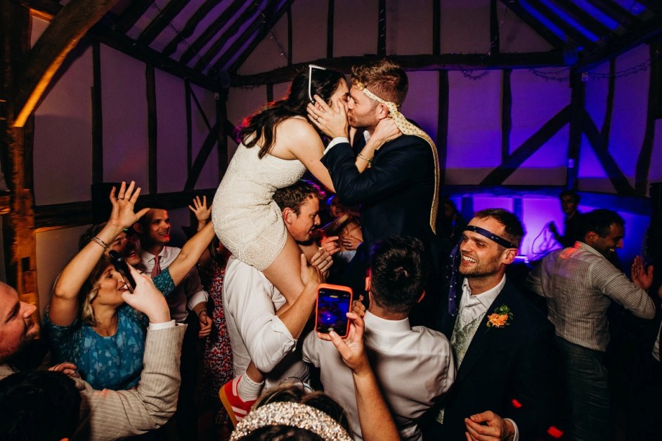 A bride in a short dress sits on a guest's shoulders, kissing the groom who is also on another