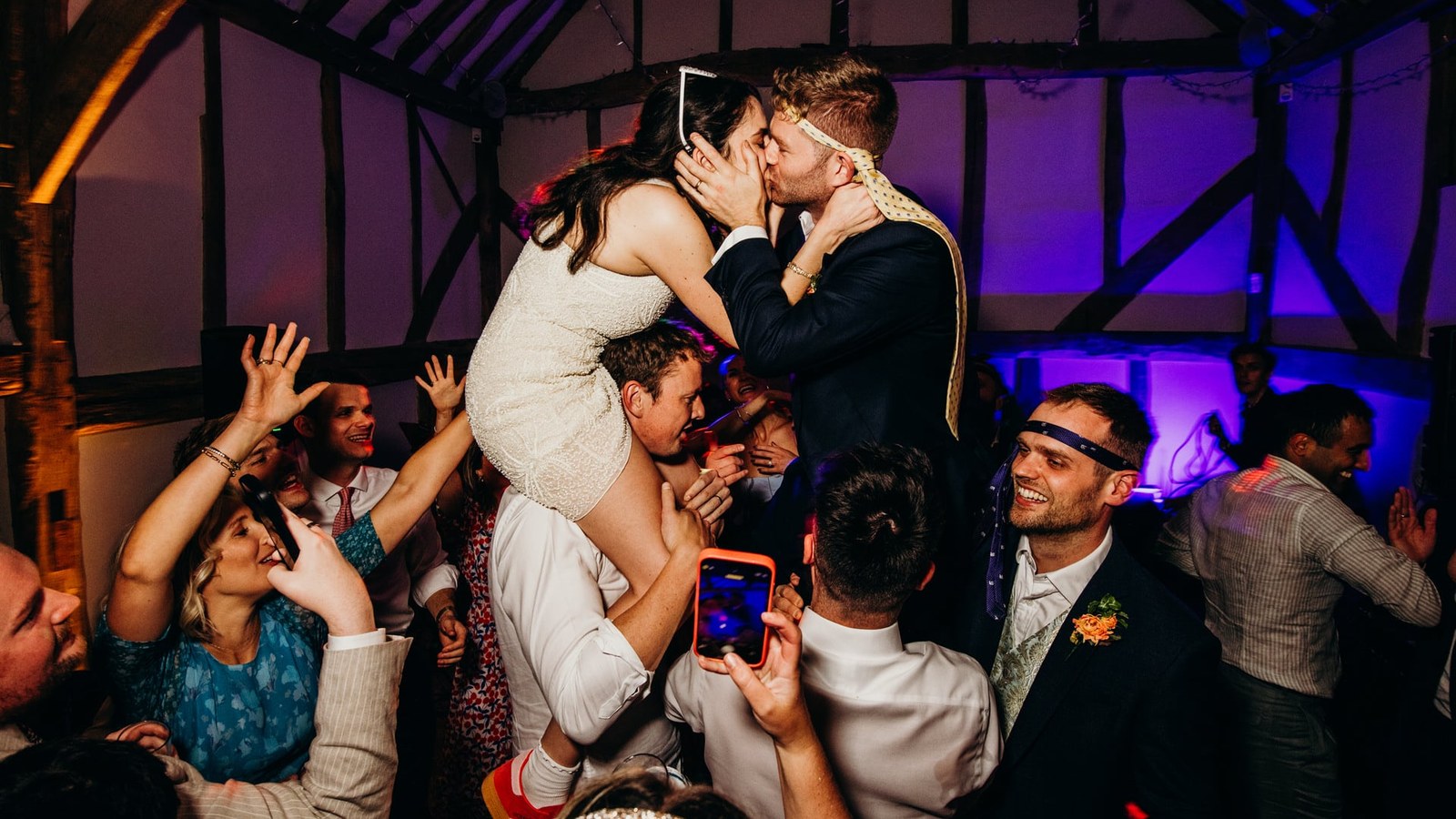 A bride in a short dress sits on a guest's shoulders, kissing the groom who is also on another