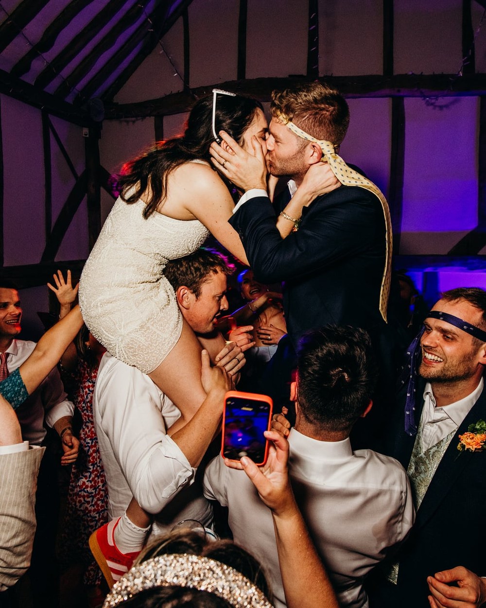 A bride in a short dress sits on a guest's shoulders, kissing the groom who is also on another