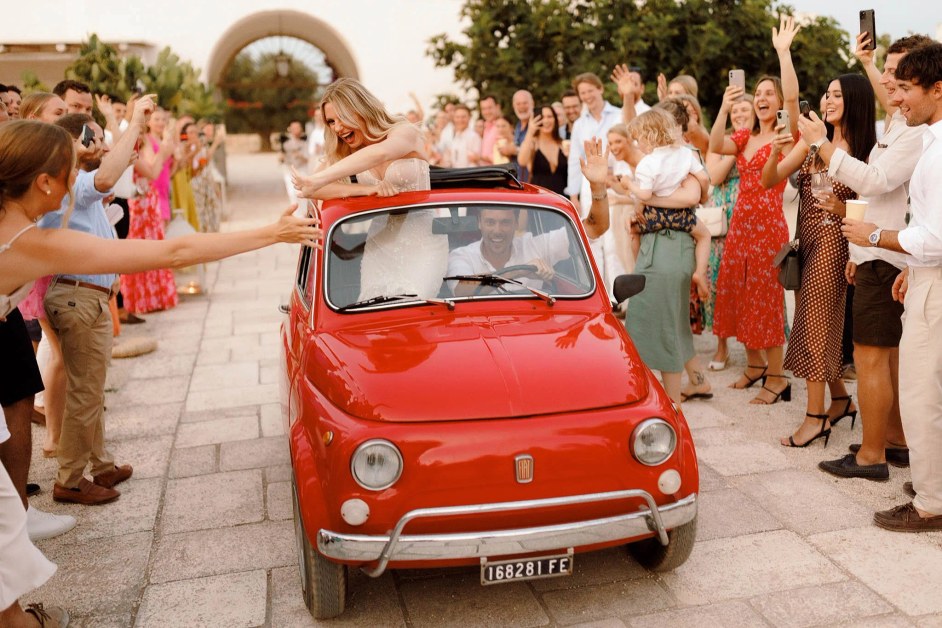 The bride, in a white strapless gown, and groom, in a white shirt, drive through a crowd of