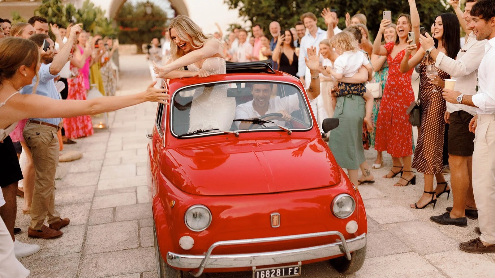 The bride, in a white strapless gown, and groom, in a white shirt, drive through a crowd of