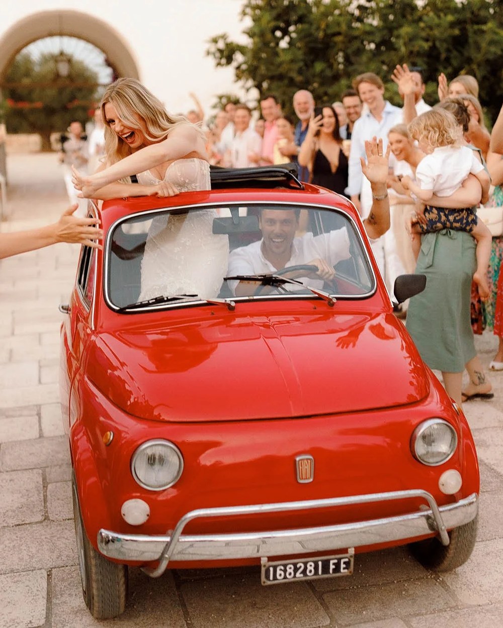 The bride, in a white strapless gown, and groom, in a white shirt, drive through a crowd of