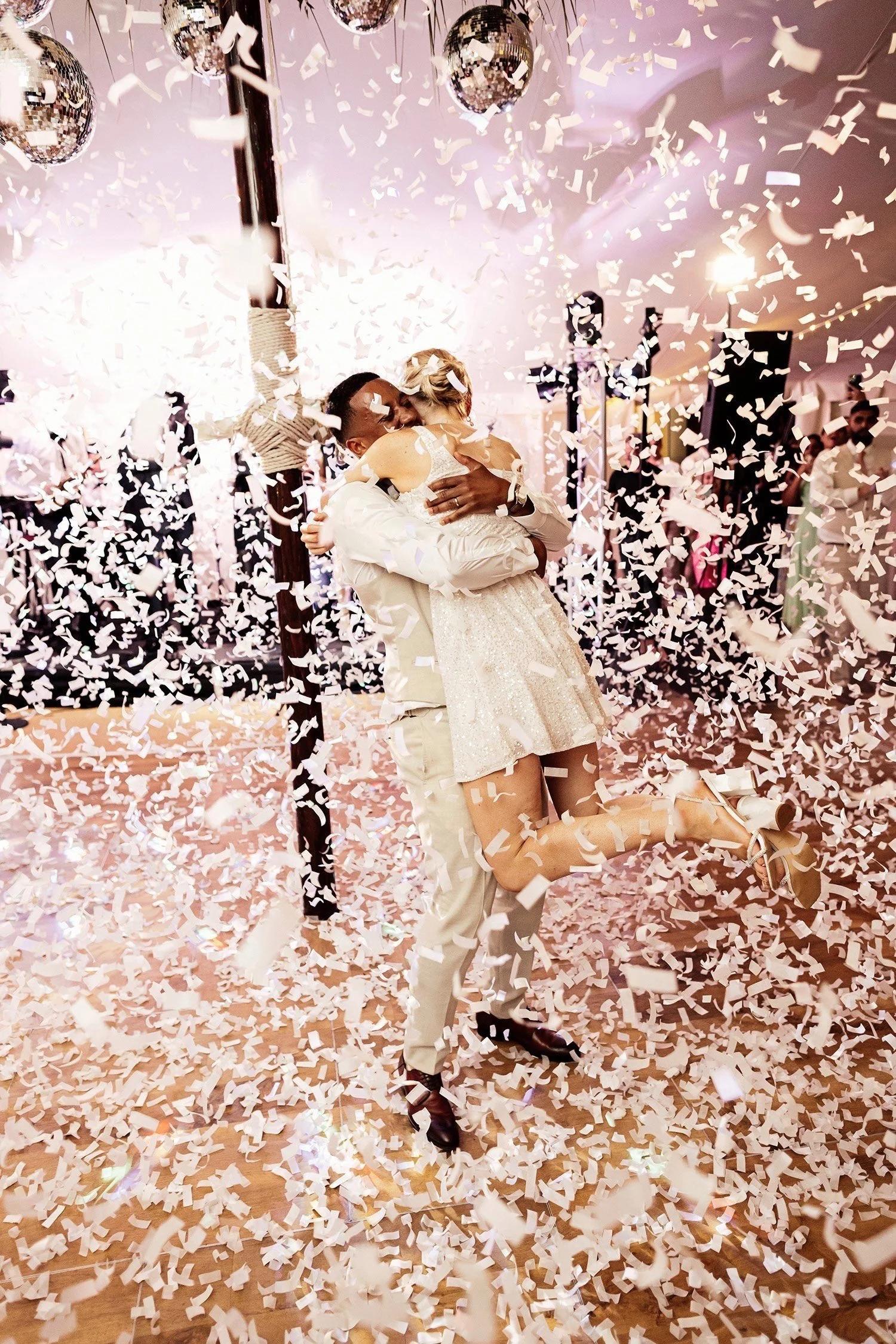 A couple embraces tightly on a wooden dance floor, surrounded by white falling confetti and disco