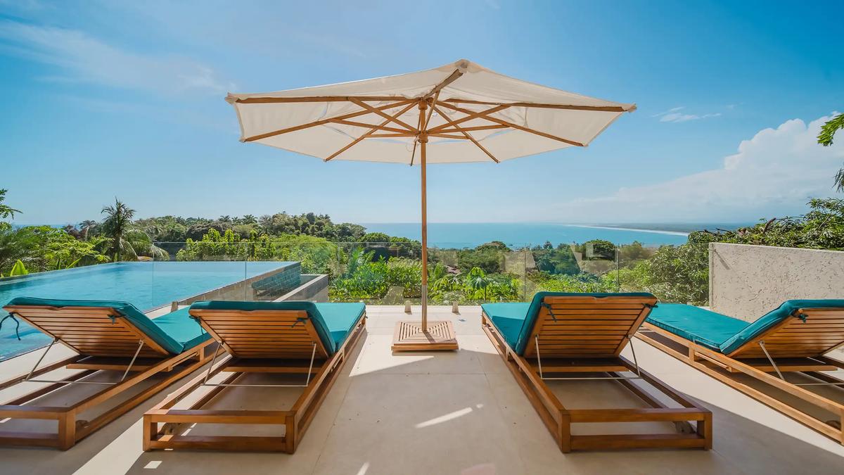 Poolside loungers under a white umbrella at Casa Nola with ocean and beach views
