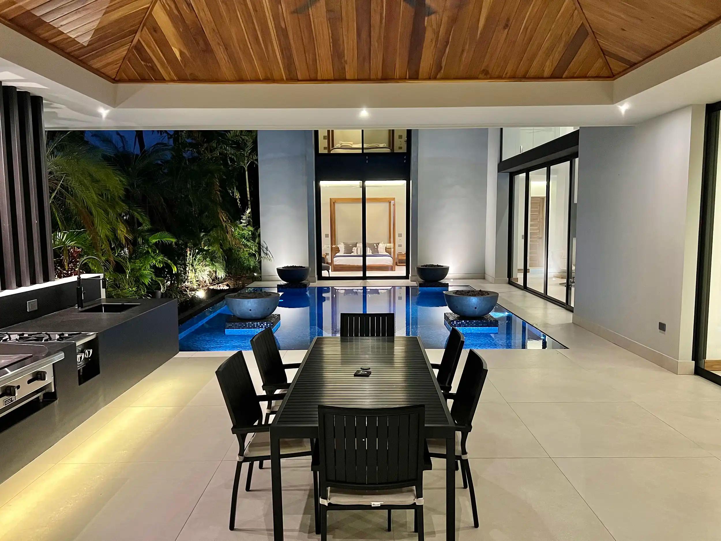 Outdoor dining table beside the lit pool and kitchen at Villa de Grace