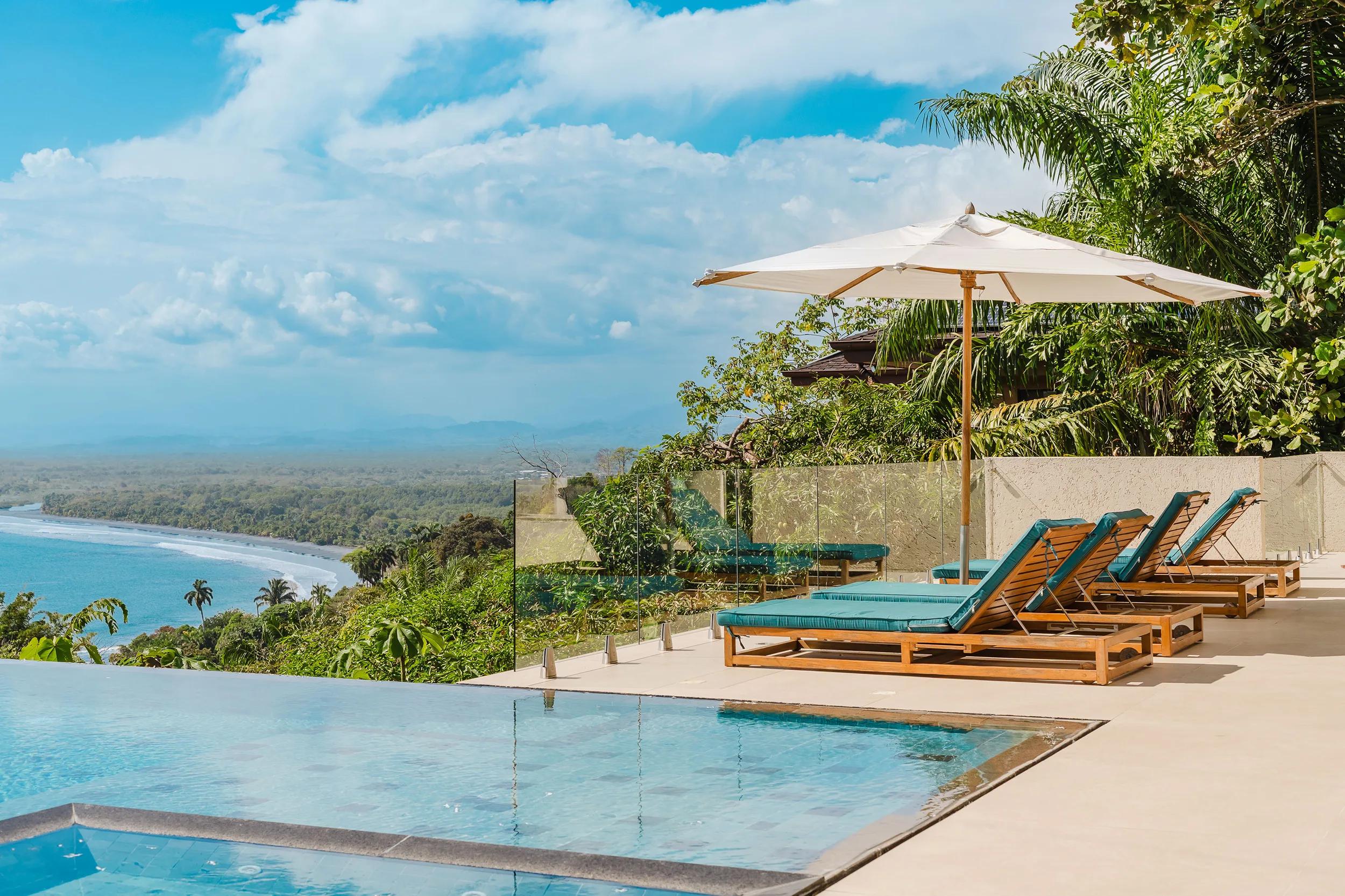 Infinity pool, loungers, and umbrella at Casa Nola with the coastline below