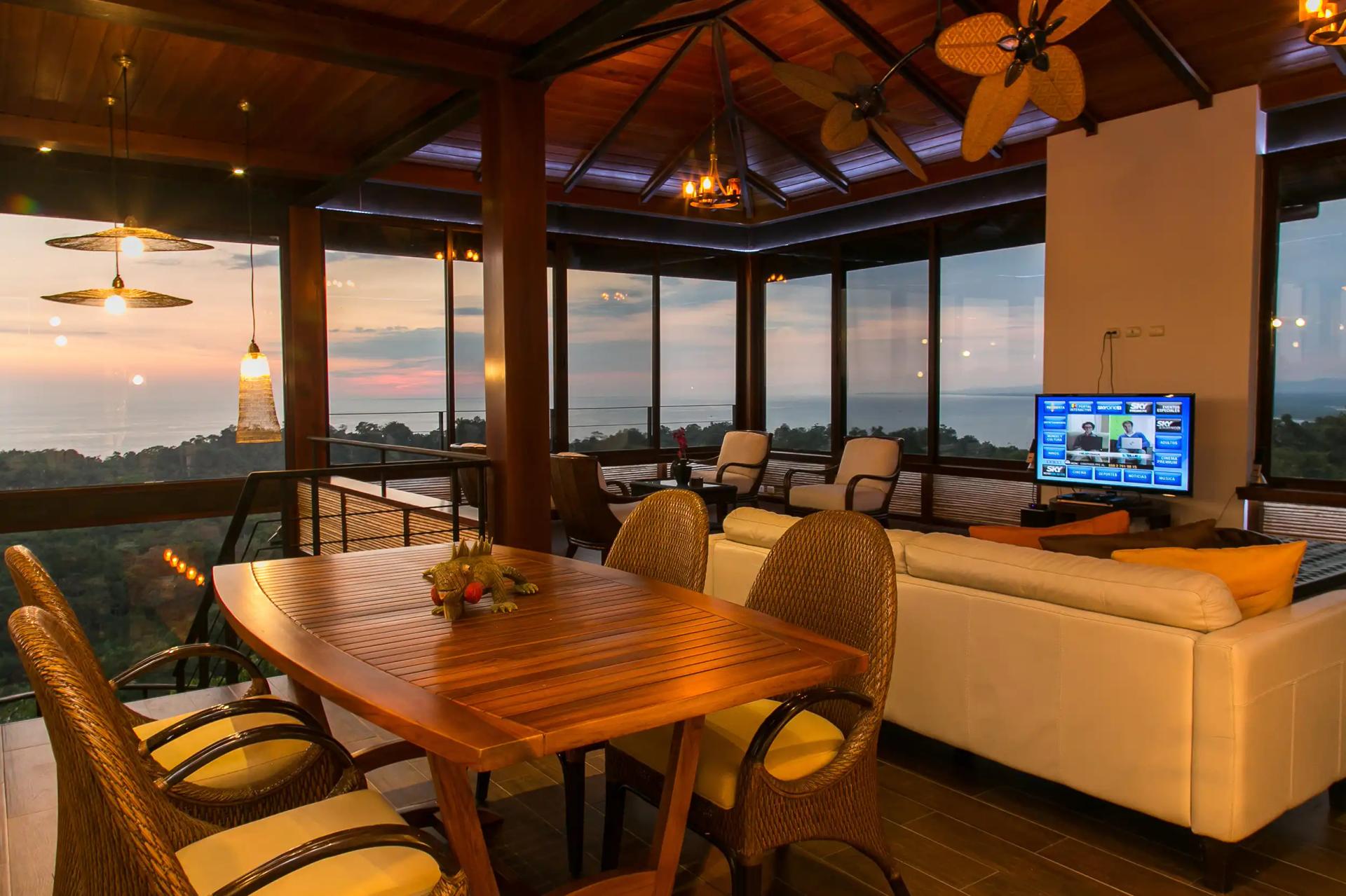 Top-floor lounge at Villa Celaje with teak dining table and wraparound sunset ocean windows