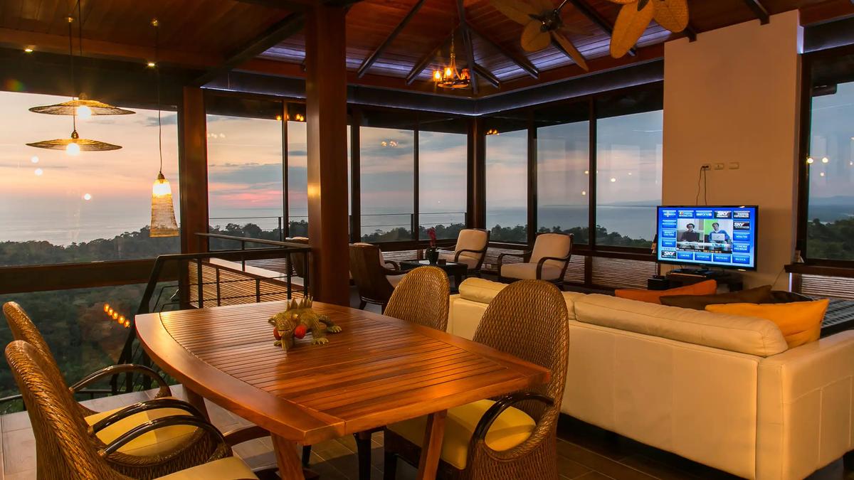 Top-floor lounge at Villa Celaje with teak dining table and wraparound sunset ocean windows