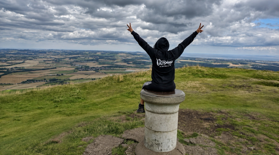 An Outplayer on a hilltop above Dundee, arms outstretched over the fields below.