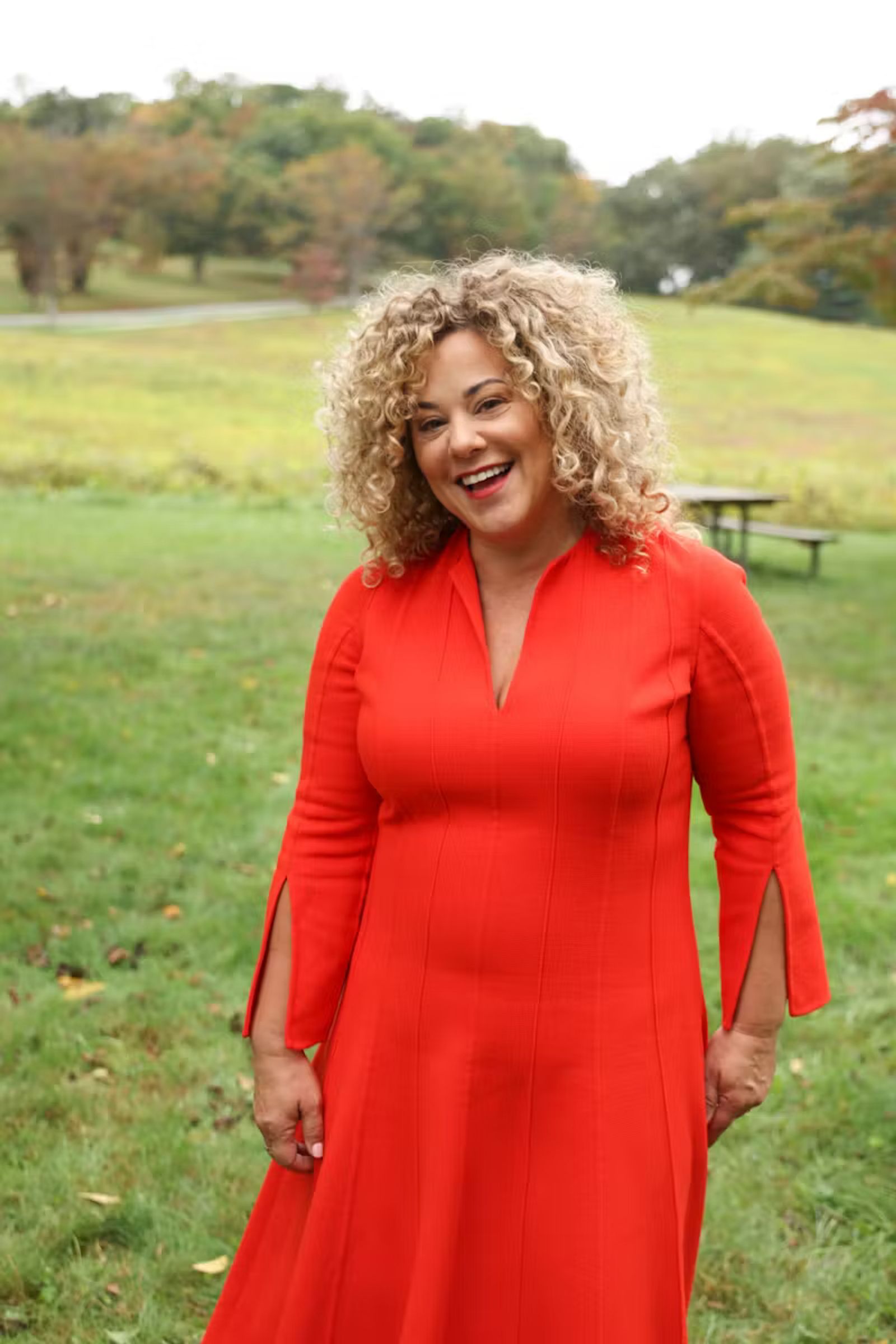 A woman with curly hair and bright red dress smiling outdoors