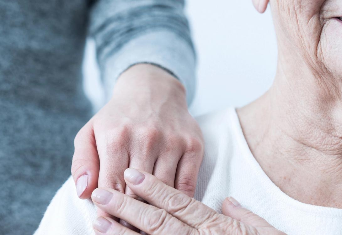 A man places his hand on an elderly woman's shoulder, the image is cropped to focus on the hand and shoulder