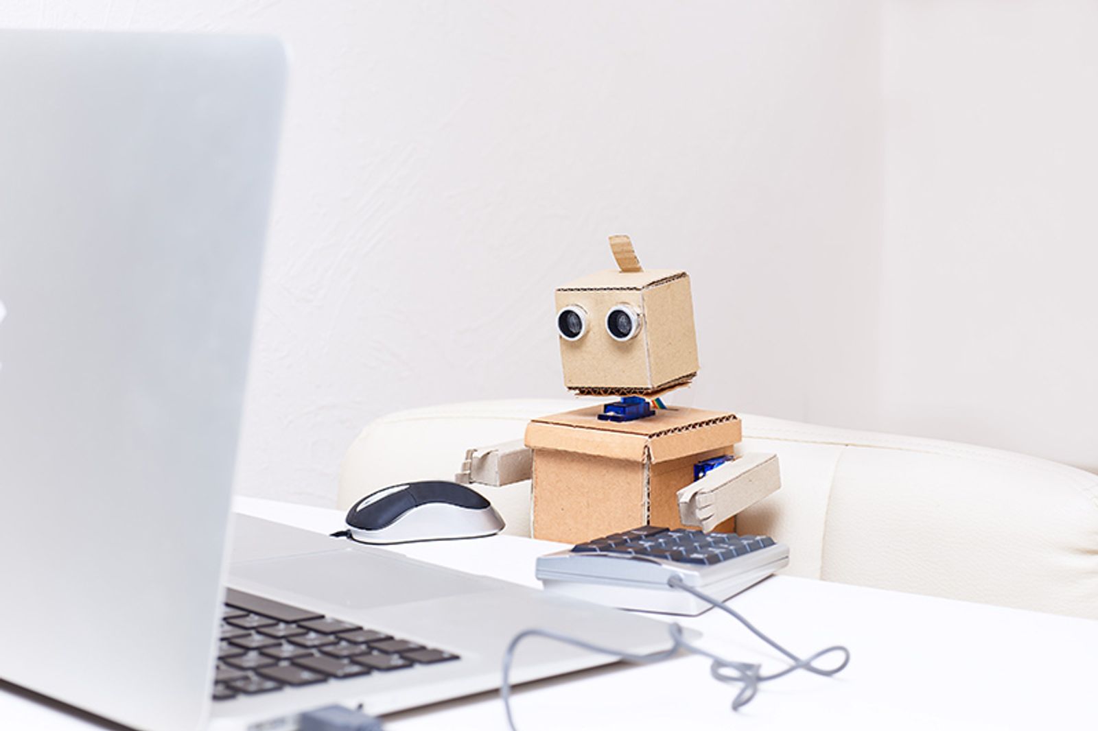 Small cardboard robot sitting at a bright desk beside a laptop, mouse, and keyboard