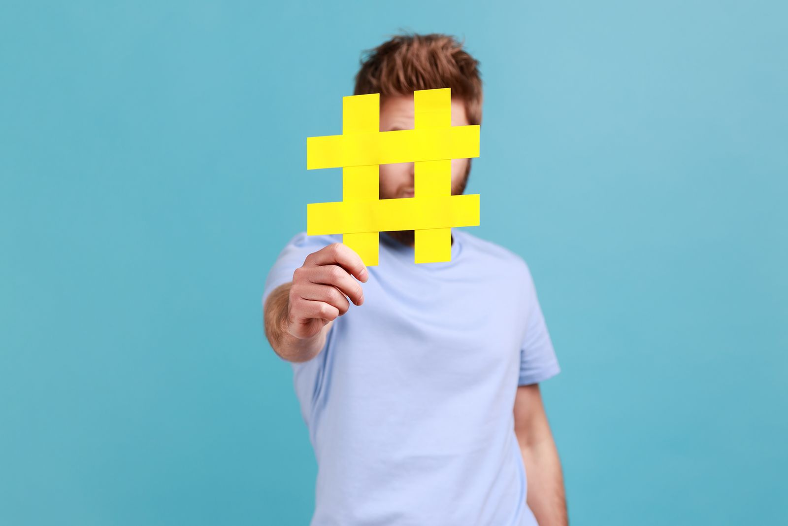 Man holding a large yellow hashtag cutout in front of his face against a blue background