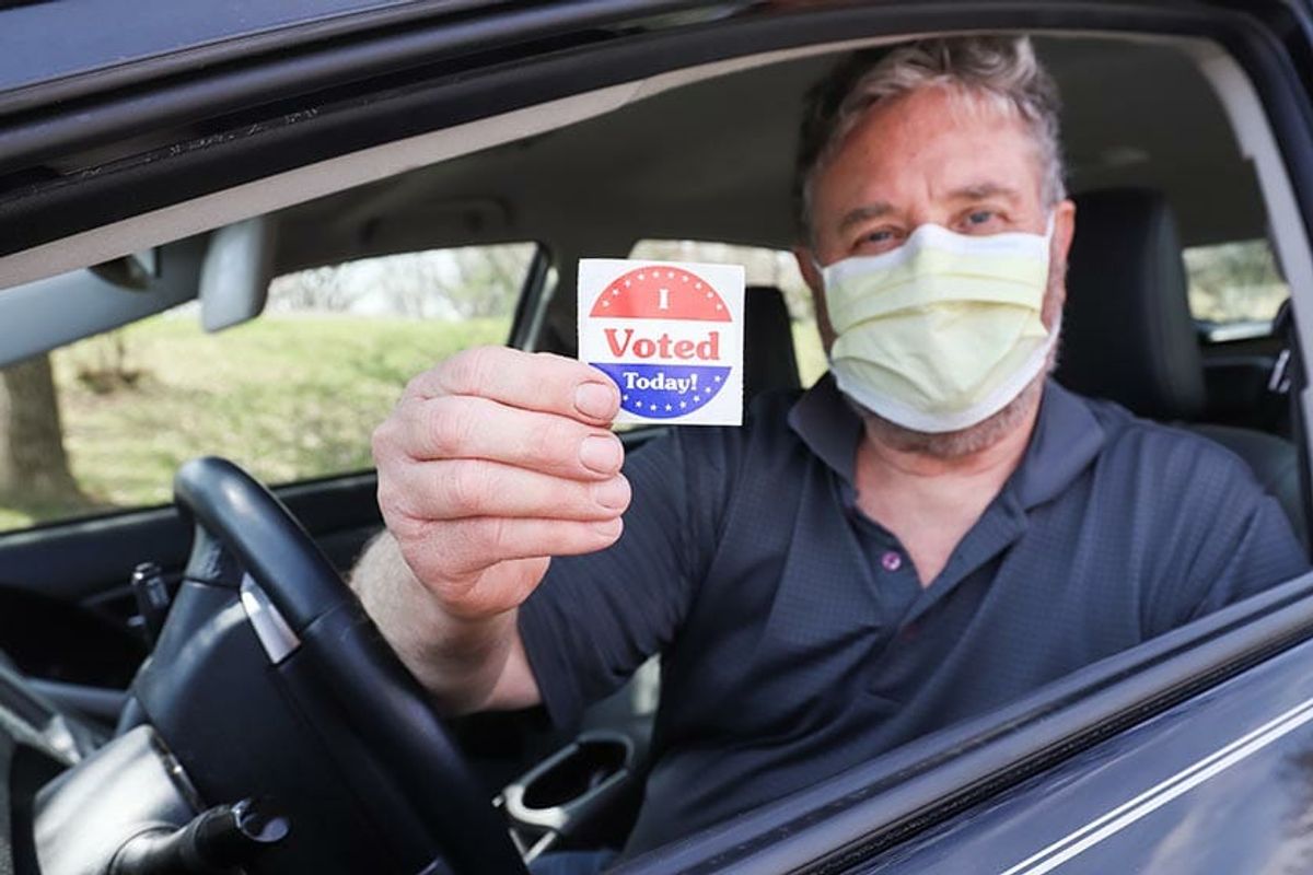 Masked voter in a car holding an I Voted Today sticker