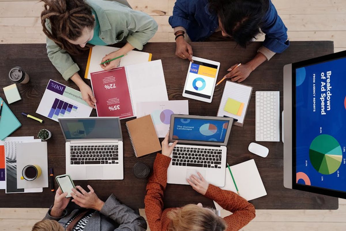 Overhead view of a media team reviewing ad spend charts across laptops, tablets and printouts