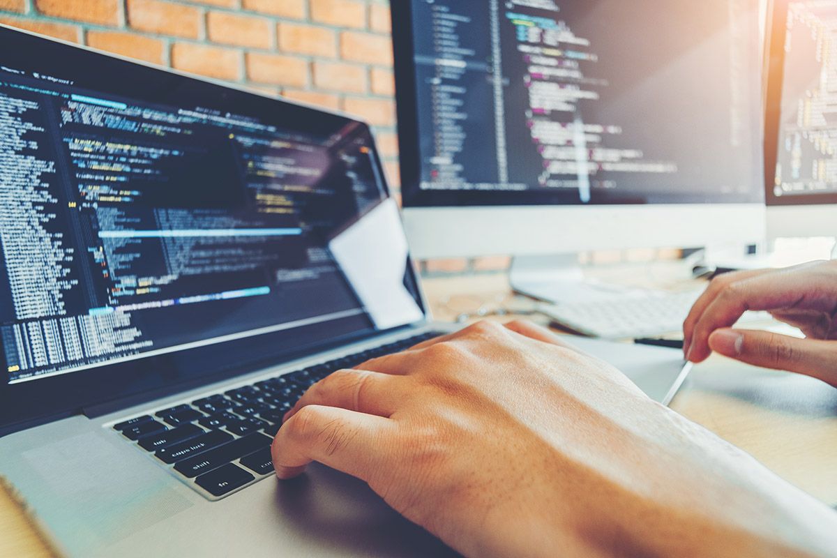 Web developer typing code on a laptop beside a monitor of source code