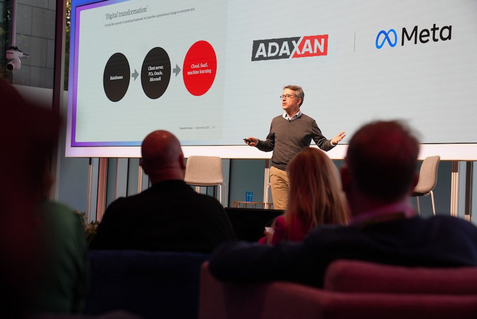 Speaker presenting on stage in front of a screen showing the Adaxan and Meta logos