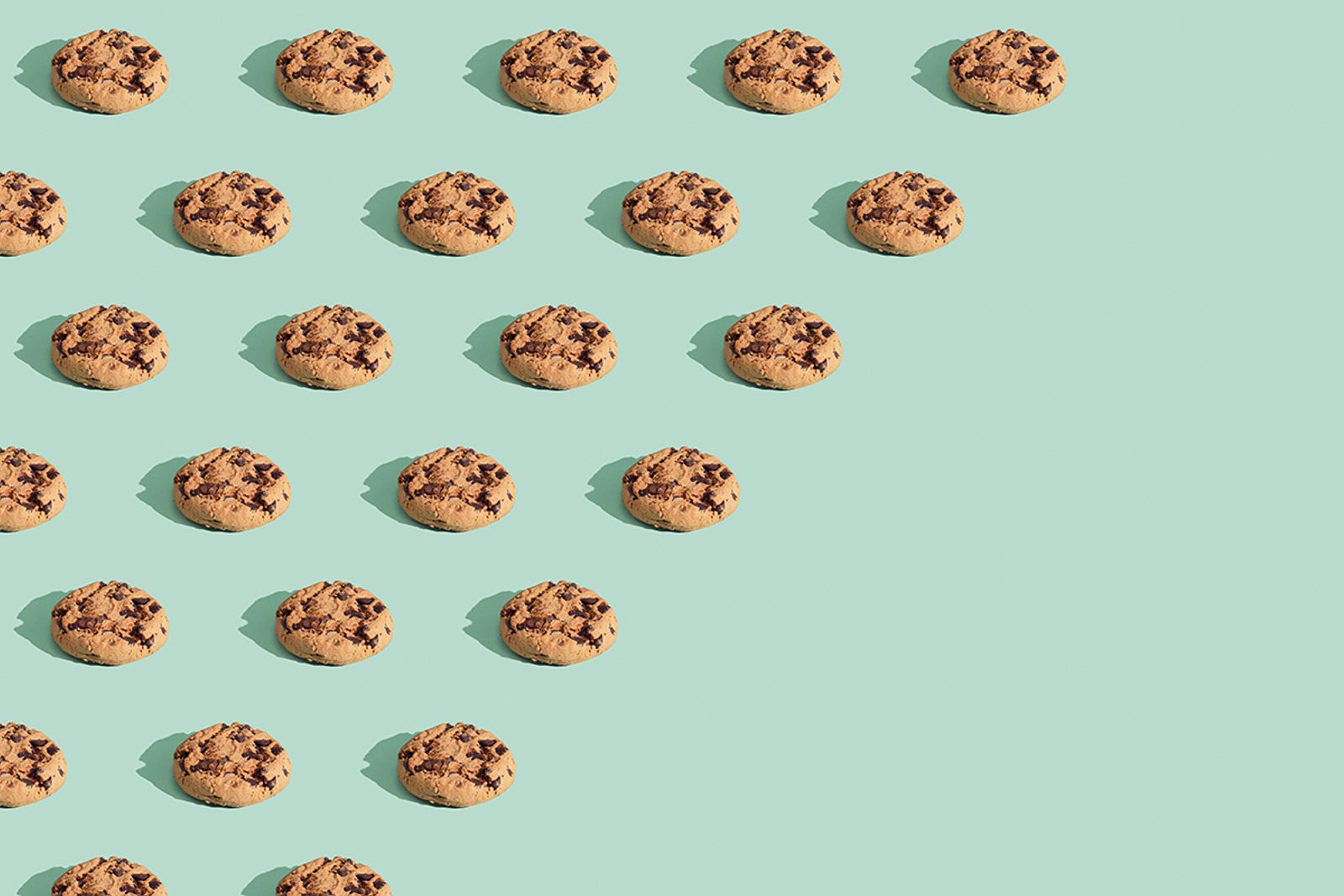 Repeating pattern of chocolate chip cookies on a mint green background fading to empty space
