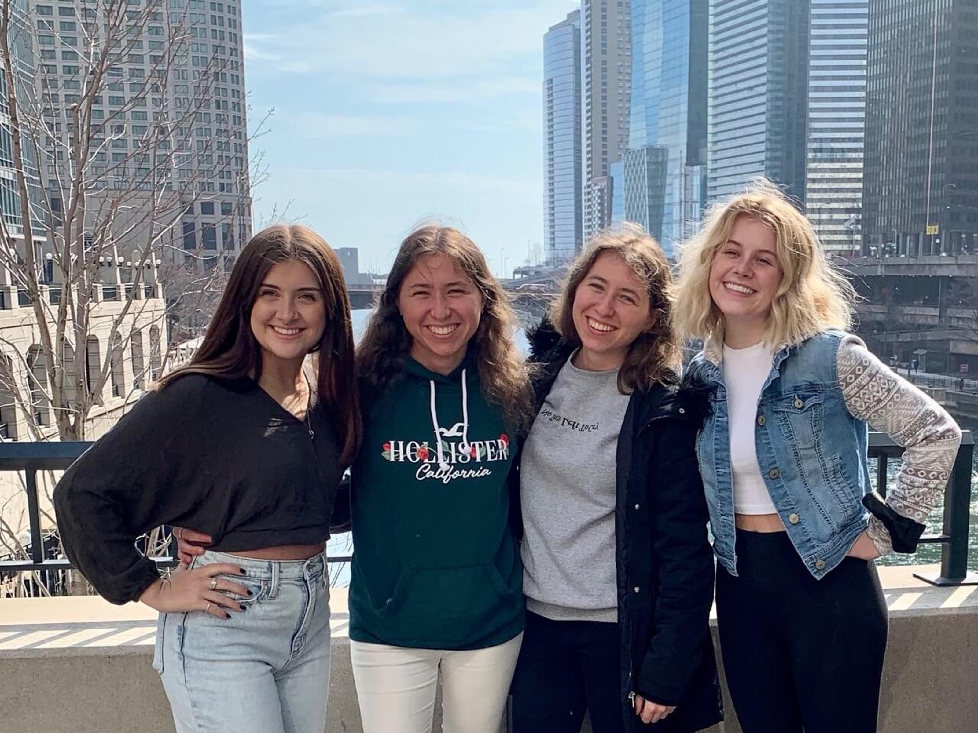 Vier junge Frauen stehen lächelnd nebeneinander auf einer Terrasse mit Blick auf eine moderne Stadtlandschaft mit Hochhäusern. Sie tragen lässige Kleidung und wirken fröhlich in der sonnigen Umgebung.