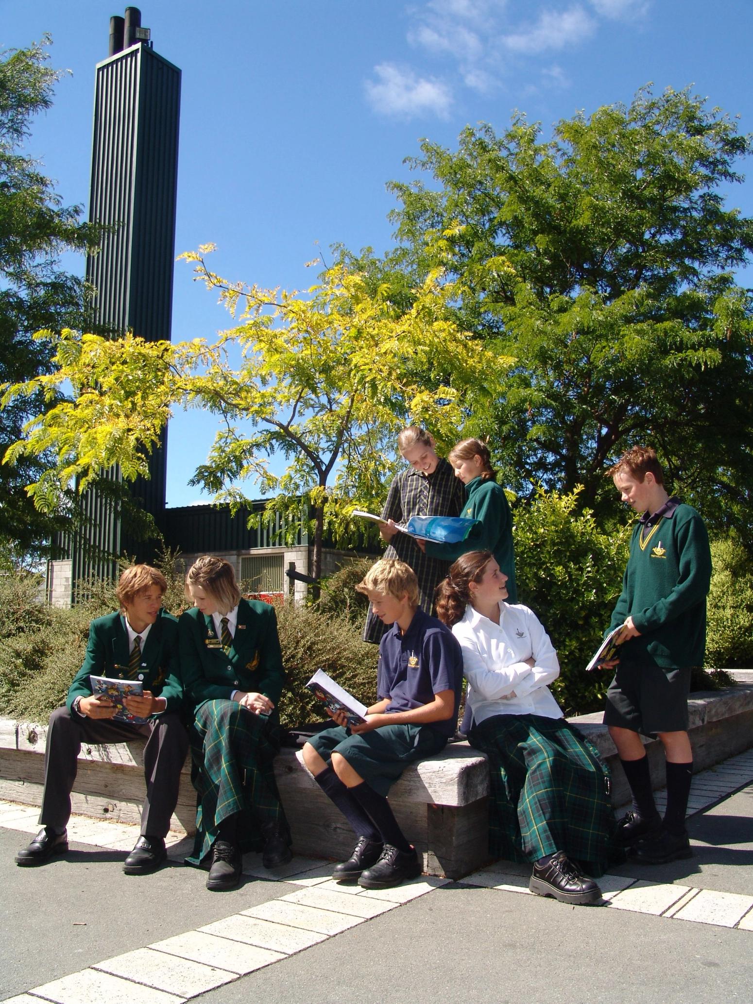Gruppe von Schülerinnen und Schülern in Schuluniformen sitzt und steht im Freien auf einer Steinmauer, liest und unterhält sich, im Hintergrund Bäume und ein moderner Turm.