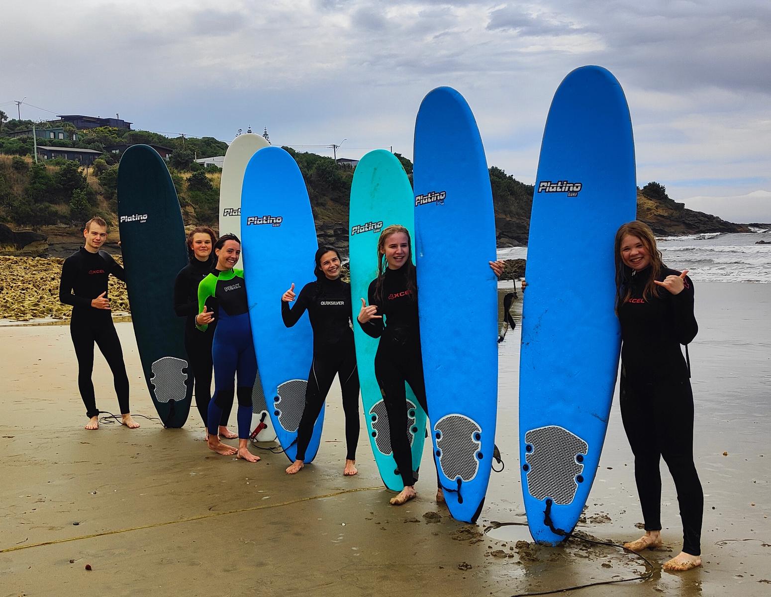 Sechs Jugendliche in Neoprenanzügen halten Surfbretter am Strand.