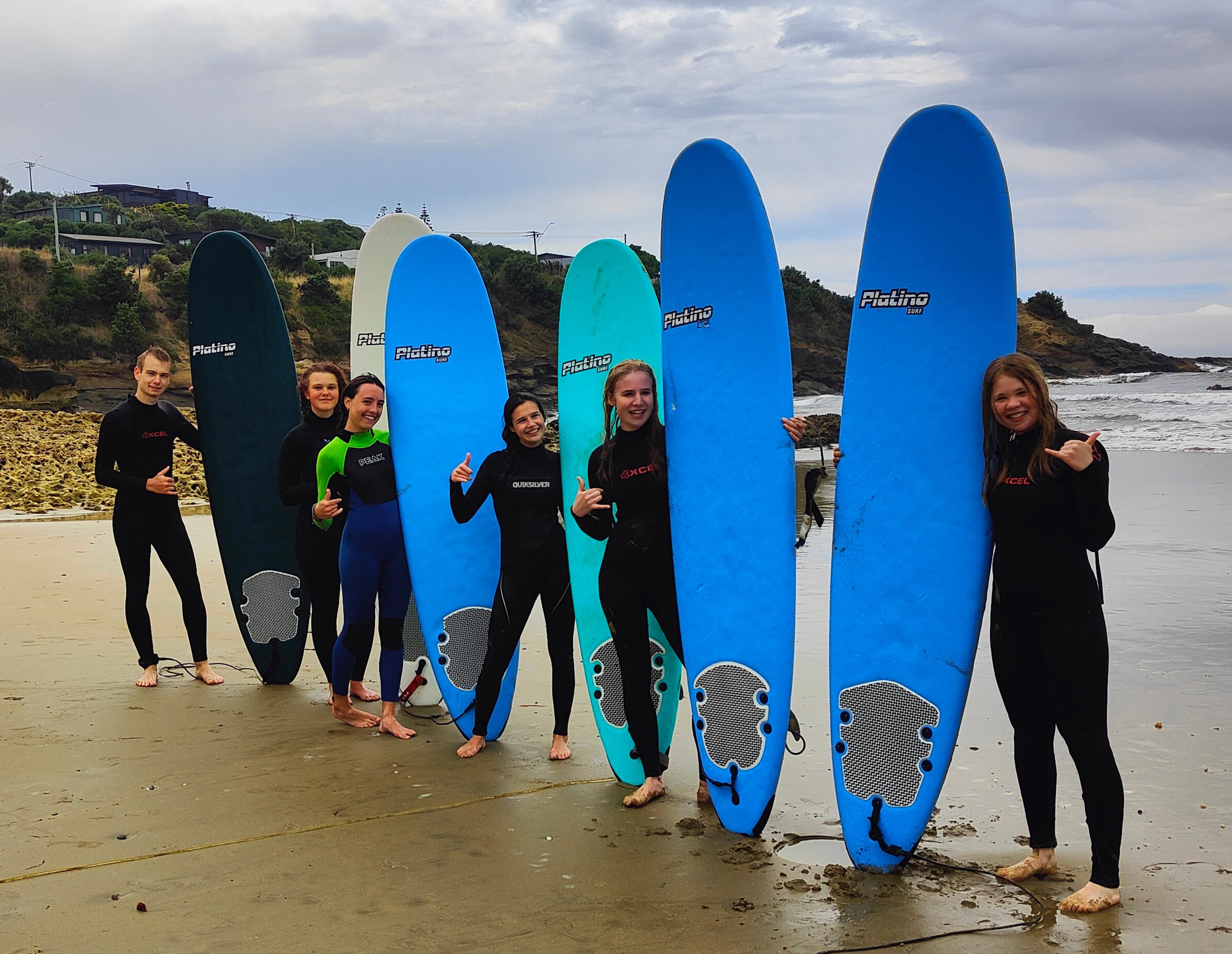 Sechs Jugendliche in Neoprenanzügen halten Surfbretter am Strand.