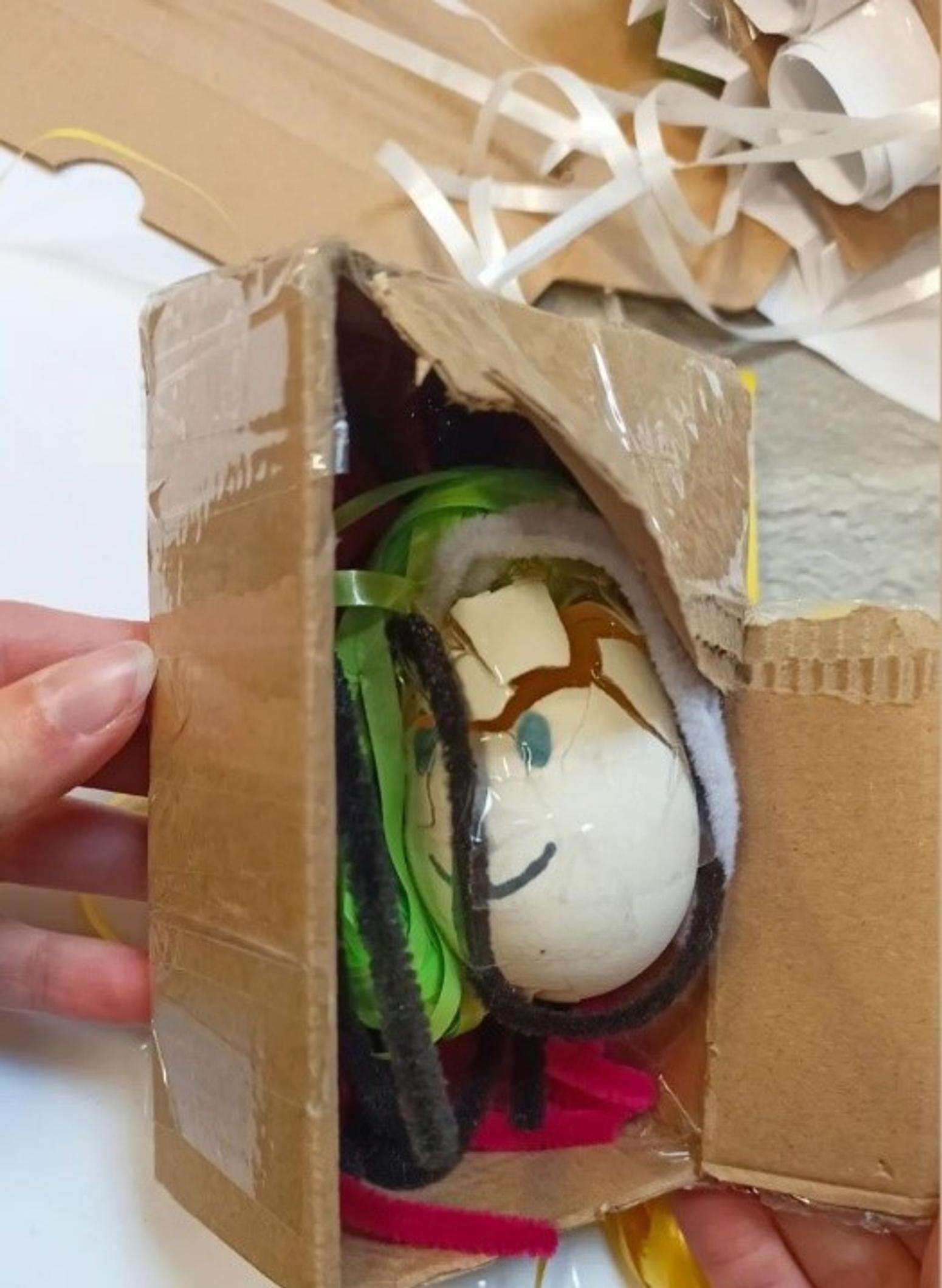 Foto von einem kaputten hartgekochten Ei in einem kleinen Pappkarton, auf dem ein Gesicht gemalt ist.
