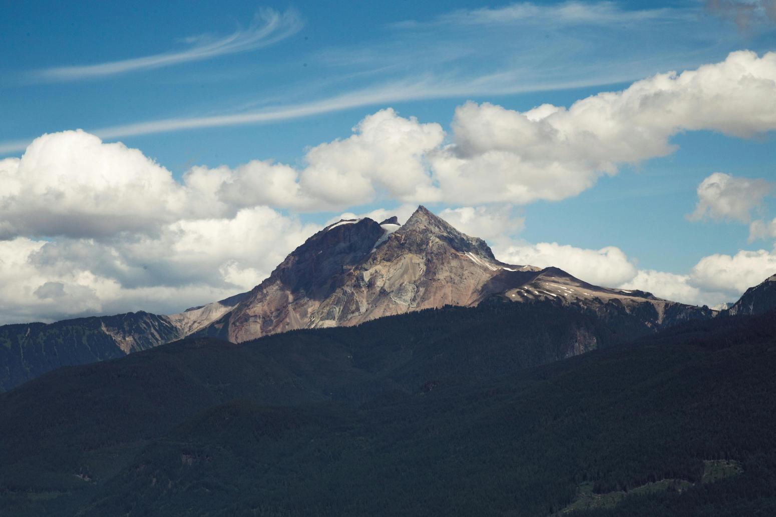 Majestätischer Berg mit bewaldeten Hängen und einer felsigen Spitze, umgeben von weißen Wolken und einem klaren blauen Himmel.